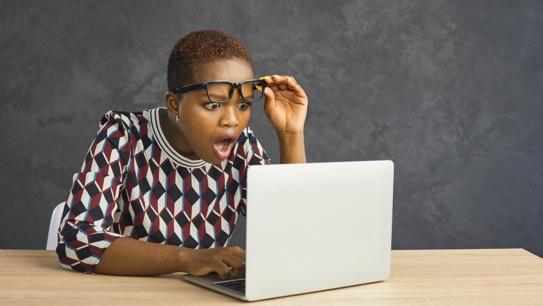 A woman lifts her glasses as she looks at a laptop screen in surprise