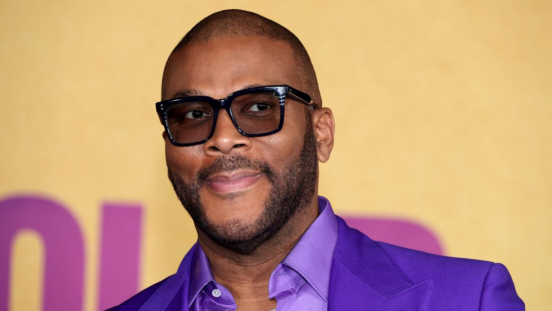 Actor Tyler Perry wears a purple jacket and lavender shirt standing before a gold background