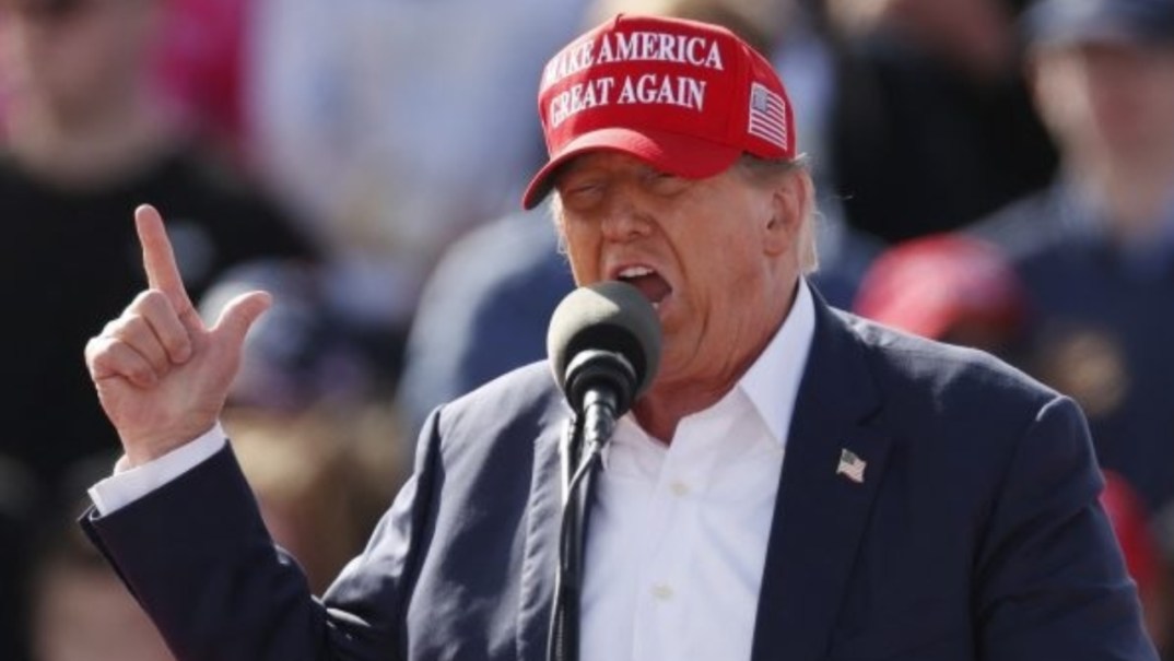 Former President Donald Trump, wearing a red MAGA hat and black jacket, points upwards as he speaks into a microphone