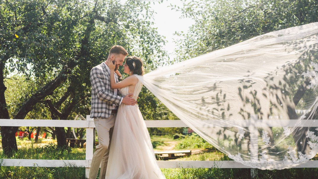 Groom and bride in flowing veil outdoors near a fence