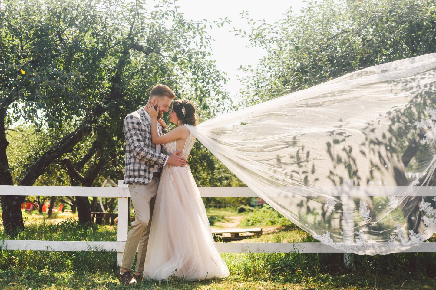 Groom and bride in flowing veil outdoors near a fence