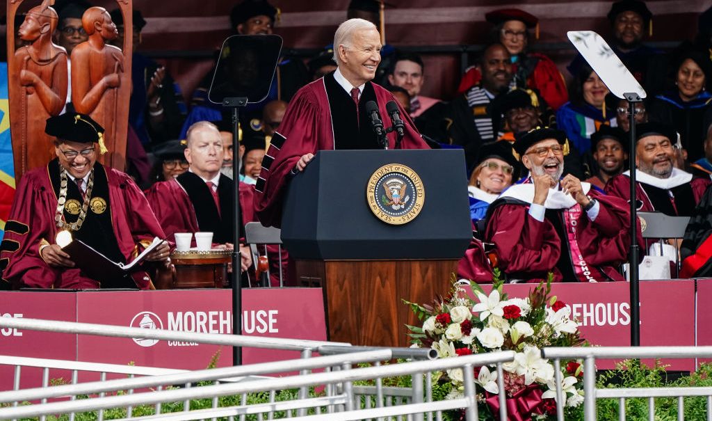 President Joe Biden stands behind a podium wearing a dark red graduate robe over a black suit and red tie