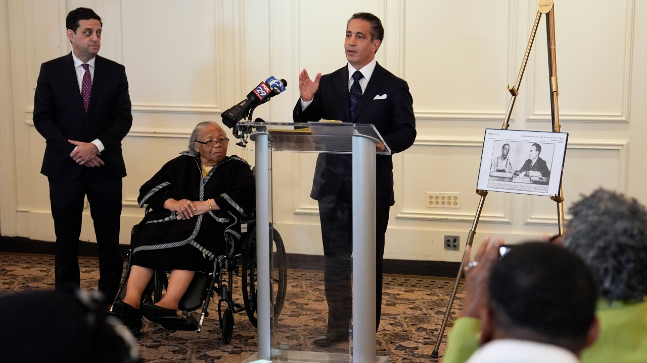 A man in a black suit speaks at a podium while another man in a suit and an older woman in a wheelchair look on